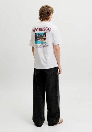 Giovane che indossa una t-shirt bianca con grafica con foto di palma a bordo piscina e testo "Negresco Nice Cote d’Azur", abbinata a jeans neri larghi.