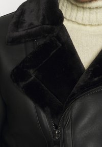 Giacca di pelle nera con colletto in peluche nero. Presenta una chiusura a zip e una superficie liscia, abbinata a un maglione crema sottostante.