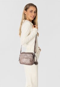 Borsa a tracolla in pelle marrone con una texture liscia, forma rettangolare, tracolla regolabile e dettagli in metallo con un ciondolo con logo.