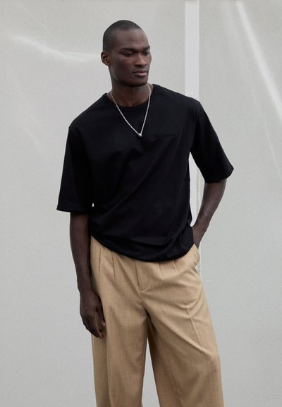 Camiseta negra de manga corta con un ajuste holgado, combinada con pantalones beige de pierna ancha con un tejido texturizado. Accesorio de collar plateado.