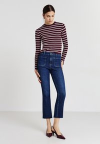 Frau, die ein burgunderfarbenes und weiß gestreiftes Langarmshirt, dunkelblaue, cropped Jeans mit Vorder-taschen und burgunderfarbene spitz zulaufende High Heels trägt.