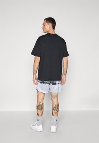 Sort kortærmet T-shirt, løs pasform, lavet af bomuldsstof. Båret sammen med mønstrede shorts og hvide sneakers, stående foran en ensfarvet baggrund.