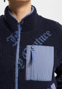 Oseba v mornarsko modri flis jopici z visokim ovratnikom, sprednjim zadrgo in svetlo modrim žepom na prsih z zadrgo.