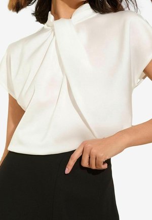 Frau trägt eine weiße kurzärmelige Bluse mit drapiertem Ausschnitt, in einen hoch taillierten schwarzen Rock gesteckt, die Hand auf der Hüfte ruhend.