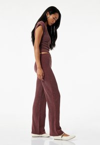 Bordeauxfarbiges, geripptes Strick-Duo bestehend aus einem enganliegenden Crop-Top und weit geschnittenen Hosen. Das Modell trägt schmal geschnittene, cremefarbene Schuhe.
