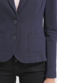 Blazer ajusté bleu marine avec deux boutons à l'avant, deux poches plaquées, porté sur un haut clair et associé à un pantalon noir.