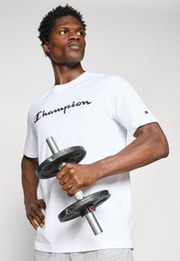 Weißes T-Shirt mit dem "Champion"-Logo in schwarzer Schreibschrift. Das Model hält eine schwarze Gewichtsscheibe mit einem silbernen Griff. Graue Shorts mit einem schlichten Design.