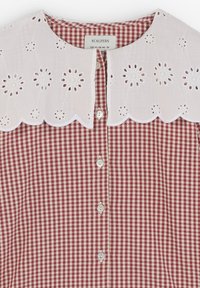 Blusa de cuadros vichy rojos y blancos con un cuello de ojal blanco con borde de pico, cierre frontal con botones y mangas cortas.