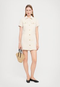 Lyhythihainen beige mekko, jossa on nappikiinnitys, kaksi rintataskua ja muotoiltu malli. Täydennetty pienellä kudotulla käsilaukulla ja mustilla tasapohjaisilla kengillä.