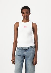 Weißes, geripptes Tanktop mit Rundhals und einem pinken, rechteckigen Grafikdesign auf der Brust; kombiniert mit hellblauen, hoch taillierten Jeans.