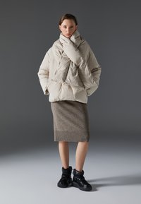 Giacca puffer beige con un design oversize e colletto ripiegato, abbinata a una gonna in maglia marrone texturizzata e sneakers platform nere.