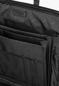 Interior de bolsa negra con cremallera que presenta múltiples compartimentos, tela texturizada y una sección de malla tejida con una etiqueta de logo discreta.