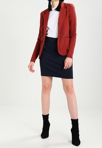 Röd skräddarsydd kavaj över en vit t-shirt, tillsammans med en marinblå pennkjol och svarta ankelstövlar. Kavajen har en slagkrage och två fickor.