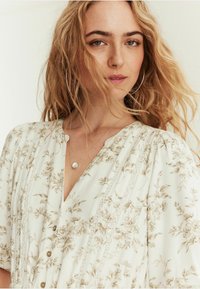 Blus med blommönster i kräm och beige med v-ringning, knappdetaljer, spetsaccent och korta ärmar. Silverhalsbandet syns.