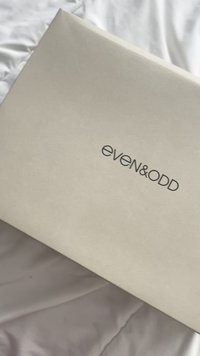 Caixa bege com o logótipo preto "even&odd" colocada sobre uma superfície de tecido branco vincado.