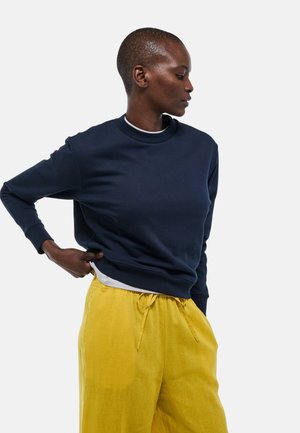 Sweat-shirt bleu marine avec un col rond et des manches longues, associé à un pantalon en lin jaune vif, ample et ceinturé à la taille.