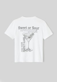 T-shirt blanc avec une impression graphique d'un verre à martini et le texte "Sweet or Sour" et "TROUVEZ VOTRE VERRE PARFAIT !" en noir.
