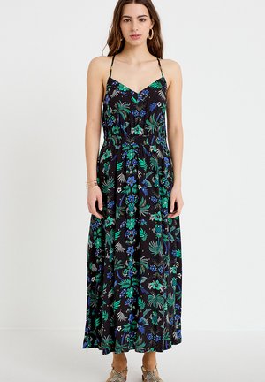 Kvinde iført en sort maxikjole med grønne og blå blomsterprint, tynde stropper og gyldne creoløreringe, stående mod en hvid baggrund.