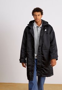 Manteau doudoune noir, long, avec une capuche, présentant une fermeture éclair et des boutons-pression, des poches latérales, et un logo blanc sur la poitrine. Porté par-dessus un polo gris.