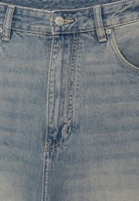 Jean en denim bleu clair en gros plan montrant un bouton, des passants de ceinture, une braguette zippée à l'avant, et une partie de la poche droite avec des détails de couture visibles.