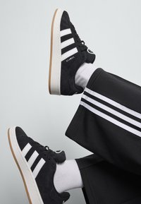 Baskets en suède noires avec un design à trois bandes blanches et un logo. Semelle en caoutchouc blanc avec une semelle extérieure en caoutchouc beige. Chaussettes blanches et pantalon noir avec des rayures blanches.