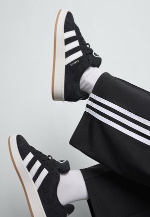 Zwarte suède sneakers met een witte drie-strepenontwerp en merknaam. Witte rubberen zool met een gumrubberen buitenzool. Witte sokken en zwarte broek met witte strepen.