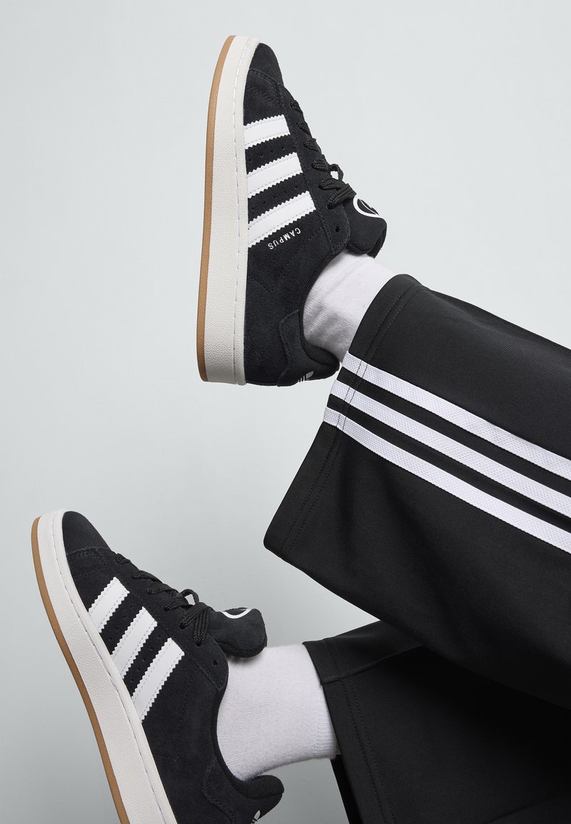Baskets en suède noires avec un design à trois bandes blanches et un logo. Semelle en caoutchouc blanc avec une semelle extérieure en caoutchouc beige. Chaussettes blanches et pantalon noir avec des rayures blanches.