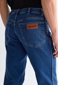 Közeli kép sötétkék Wrangler farmert viselő személyről, aki hátulnézetből mutatja a hátsó zsebeket és a jobb oldali zseb logómatricáját.