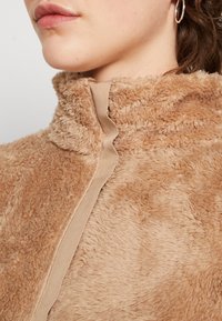 Beiger, flauschiger Stoff mit hohem Kragen und glattem, beigem Reißverschluss. Weiche Textur mit sichtbaren Plüschfasern.