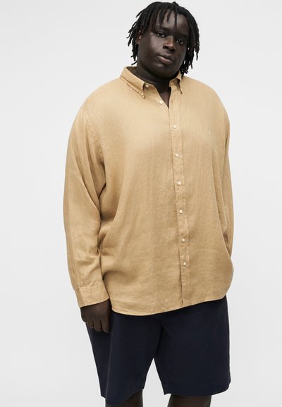 Άνδρας με σκούρο δέρμα και κοτσίδες, που φοράει άνετο μπεζ πουκάμισο με κουμπιά και μαύρο σορτς, στέκεται μπροστά σε απλό φωτεινό φόντο.