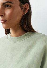 Gaiši zaļa džemperis no mīksta materiāla ar ribotu apkakli un gludu faktūru, daļēji redzams kopā ar cilvēka kaklu un seju.