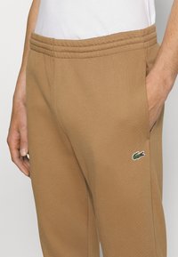 Pantaloni de trening maro din material moale, cu un design conic, talie elastică cu detalii texturate și un mic logo brodat.