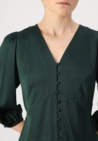 Smaragdgrønn bluse med V-hals, puffermer og knappelukking foran. Glatt stoff med subtil glans og tettsittende design.
