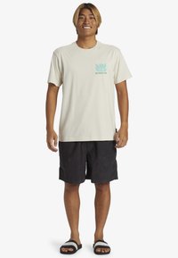 Beige bomulds t-shirt med teal grafisk design på venstre bryst, parret med sorte shorts og sorte sandaler med hvide accenter.