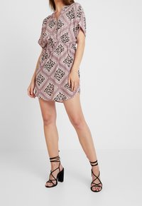 Woman wearing pink and black patterned short dress with drawstring waist and black strappy high-heeled sandals standing on white floor.