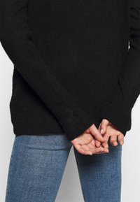 Pull en maille noir à coupe décontractée, avec des poignets côtelés et une surface texturée, porté avec un jean bleu ajusté.