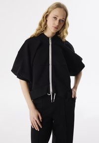 Zwarte cropped sweatshirt met brede mouwen, rits aan de voorkant en een trekkoord aan de onderkant. Zachte stof met een gladde textuur en een casual pasvorm.