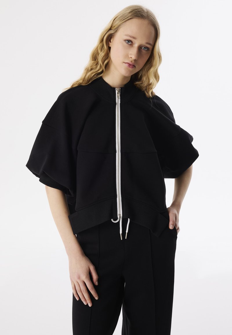 Zwarte cropped sweatshirt met brede mouwen, rits aan de voorkant en een trekkoord aan de onderkant. Zachte stof met een gladde textuur en een casual pasvorm.