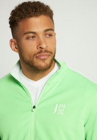 Lysgrøn zip-up jakke med høj krave, hvide detaljer og logo-design på brystet. Glat stof med en tætsiddende silhuet.