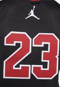 Svart baskettröja med röd och vit siffra "23" på baksidan, med en vit Jumpman-logotyp högst upp. Tillverkad av vadderad nätmaterial.