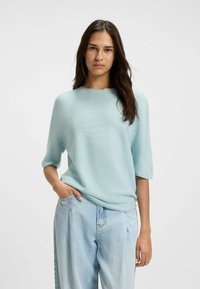 Maglione lavorato a maglia blu chiaro con maniche corte, scollo rotondo e tessuto texturizzato. Indossato sopra jeans blu chiaro a vestibilità rilassata, mani nelle tasche.