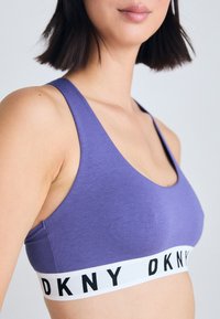 Lilla sports-bh med en bred hvit kant som viser "DKNY" i svart. Myk stoff, racerback-design og sømløs tekstur.
