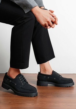 Personne assise, mains jointes, portant un pantalon noir raccourci et des chaussures noires lacées avec des semelles épaisses sur un sol en bois.