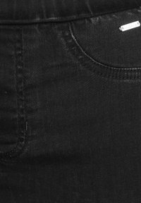 Crni traper tkanina izbliza, s prednjim džepom i suptilnim sjajem. Uključuje detalje šavova i mali metalni logotip.