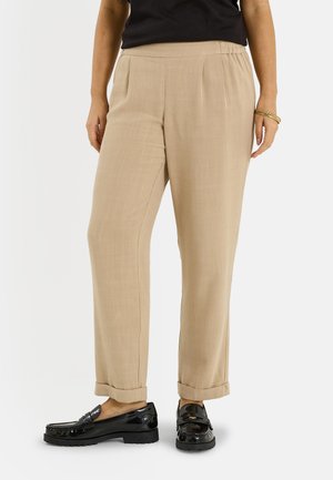 Pantalon beige à coupe ample avec ourlets retroussés, porté avec des mocassins en cuir verni noir et un haut noir, bracelets dorés à un poignet.