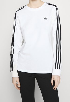 Osoba v bílém dlouhém tričku Adidas s černými třemi pruhy na rukávech a černých kalhotách, stojící před jednobarevným pozadím.