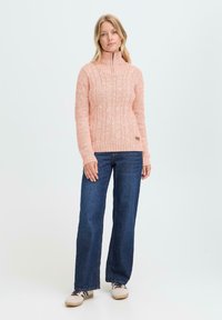 Kvinne som står i en rosa kabelstrikkegenser med glidelås krage, blå vide jeans og beige sneakers mot en enkel lyse bakgrunn.
