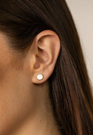 Gros plan sur l'oreille d'une femme aux cheveux bruns foncés et raides portant une petite boucle d'oreille clou ronde en or et nacre blanche.