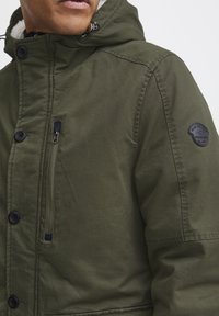Chaqueta verde oliva con un bolsillo en el pecho con cremallera, cierre de gran botón y capucha forrada de piel. Presenta un parche de la marca en la manga.
