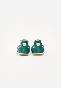 Grüne Wildleder-Sportschuhe mit weißen Akzenten, runden Formen, flachen Gummisohlen und einem kleinen gelben Logo an der Rückseite der Ferse.